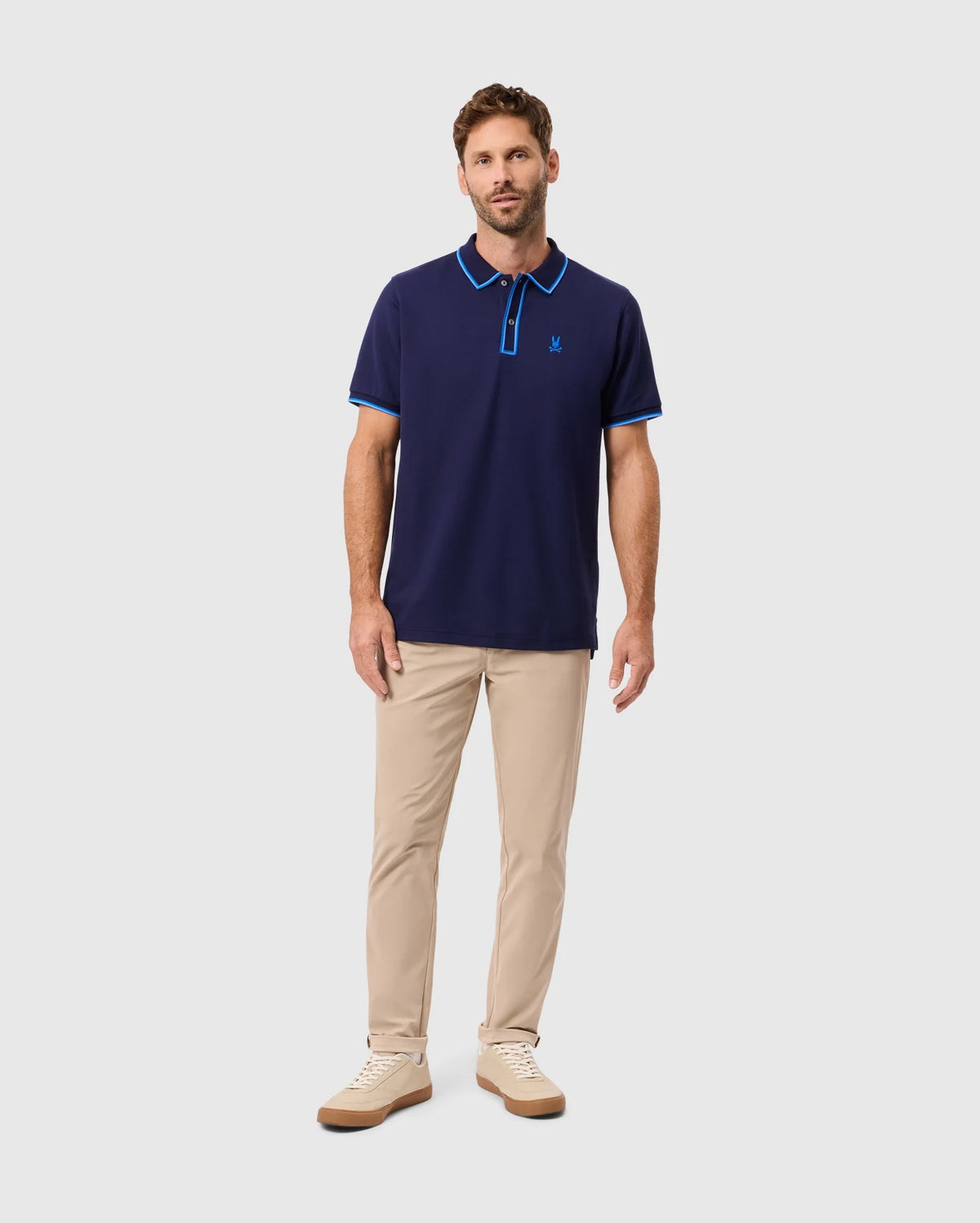 A man stands wearing the stylish Psycho Bunny Men's Kent Pique Polo Shirt, B6K951D200, in navy with blue accents, paired with beige pants and sneakers against a light gray background.