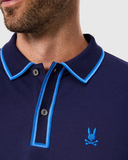 The Psycho Bunny Men's Kent Pique Polo Shirt (B6K951D200) features a stylish close-up look with dark blue fabric, light blue trim and buttons, and an embroidered rabbit emblem above crossed bones on the chest, blending comfort and flair.