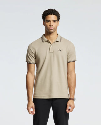 Wearing a Psycho Bunny MENS MONROE PIQUE POLO - B6K964D200 in beige with black accents, Mother-of-Pearl buttons, and a small embroidered logo on the chest, the person stands against a plain background. The shirt perfectly complements their short, curly hair as they look directly at the camera.