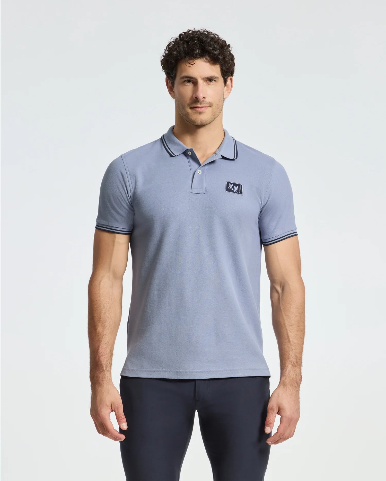 A man stands against a white background wearing a Psycho Bunny MENS MONROE PIQUE POLO - B6K964D200, featuring short sleeves and a logo appliqué on the chest. His dark pants and curly hair complete his relaxed look, with arms resting calmly by his sides.