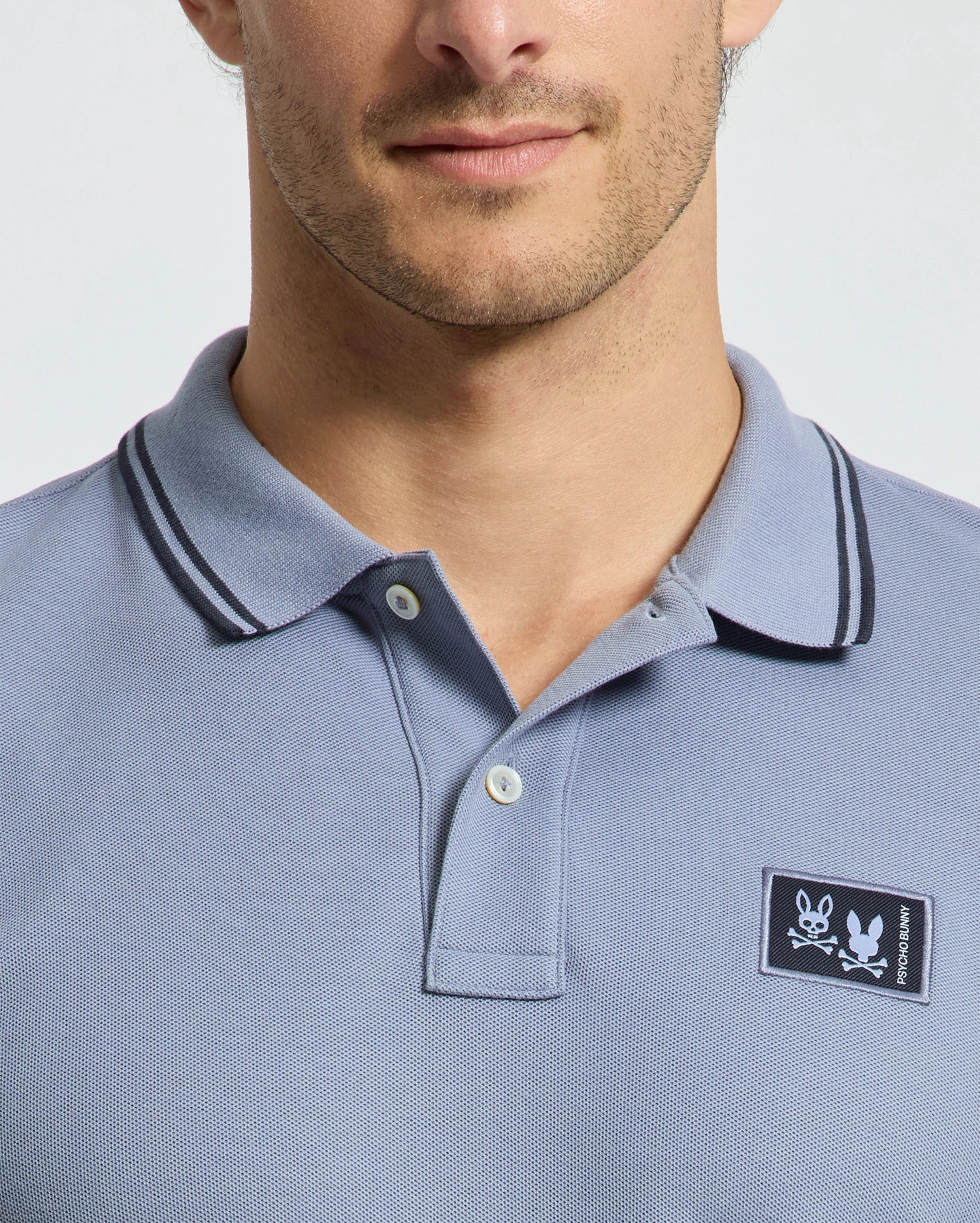 A man is seen from the shoulders up wearing a light blue Psycho Bunny MENS MONROE PIQUE POLO (B6K964D200) with dark collar trim, a logo appliqué patch on the chest, and a button-up collar in a regular fit.