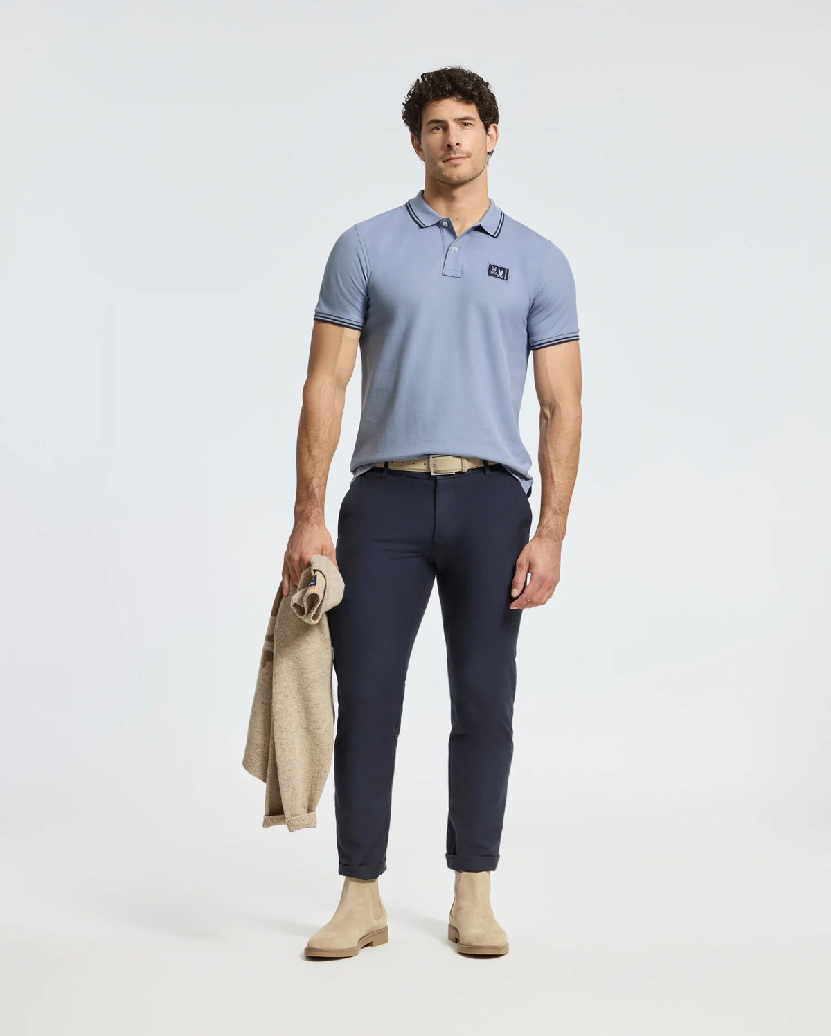 A man stands against a plain background wearing a light blue MENS MONROE PIQUE POLO by Psycho Bunny, paired with dark blue pants and beige shoes. Holding a light brown jacket, his neutral expression complements the polo's regular fit and subtle logo appliqué.