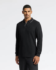 Against a plain background, a person models the Psycho Bunny MENS CREEDE PIQUE JOHNNY COLLAR POLO - B6M265E200. The black Pima cotton long sleeve shirt features a Johnny collar and white trim. The person gazes directly at the camera with a relaxed expression.