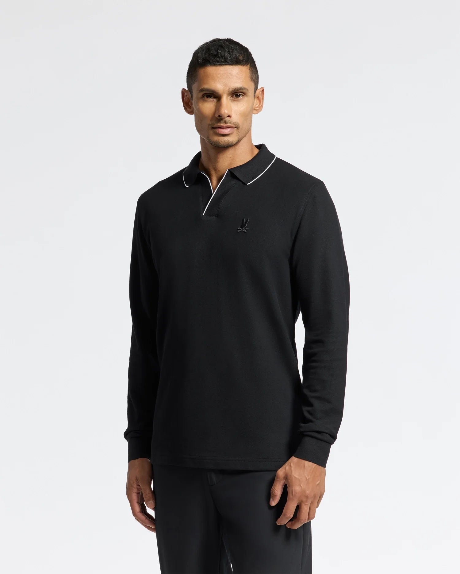 Against a plain background, a person models the Psycho Bunny MENS CREEDE PIQUE JOHNNY COLLAR POLO - B6M265E200. The black Pima cotton long sleeve shirt features a Johnny collar and white trim. The person gazes directly at the camera with a relaxed expression.