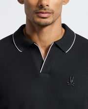 A man is wearing the MENS CREEDE PIQUE JOHNNY COLLAR POLO - B6M265E200 by Psycho Bunny. This black long sleeve polo features a Johnny collar and white piping, crafted from Pima cotton. It includes an embroidered logo of a bunny with crossbones on the chest against a plain light-colored background.