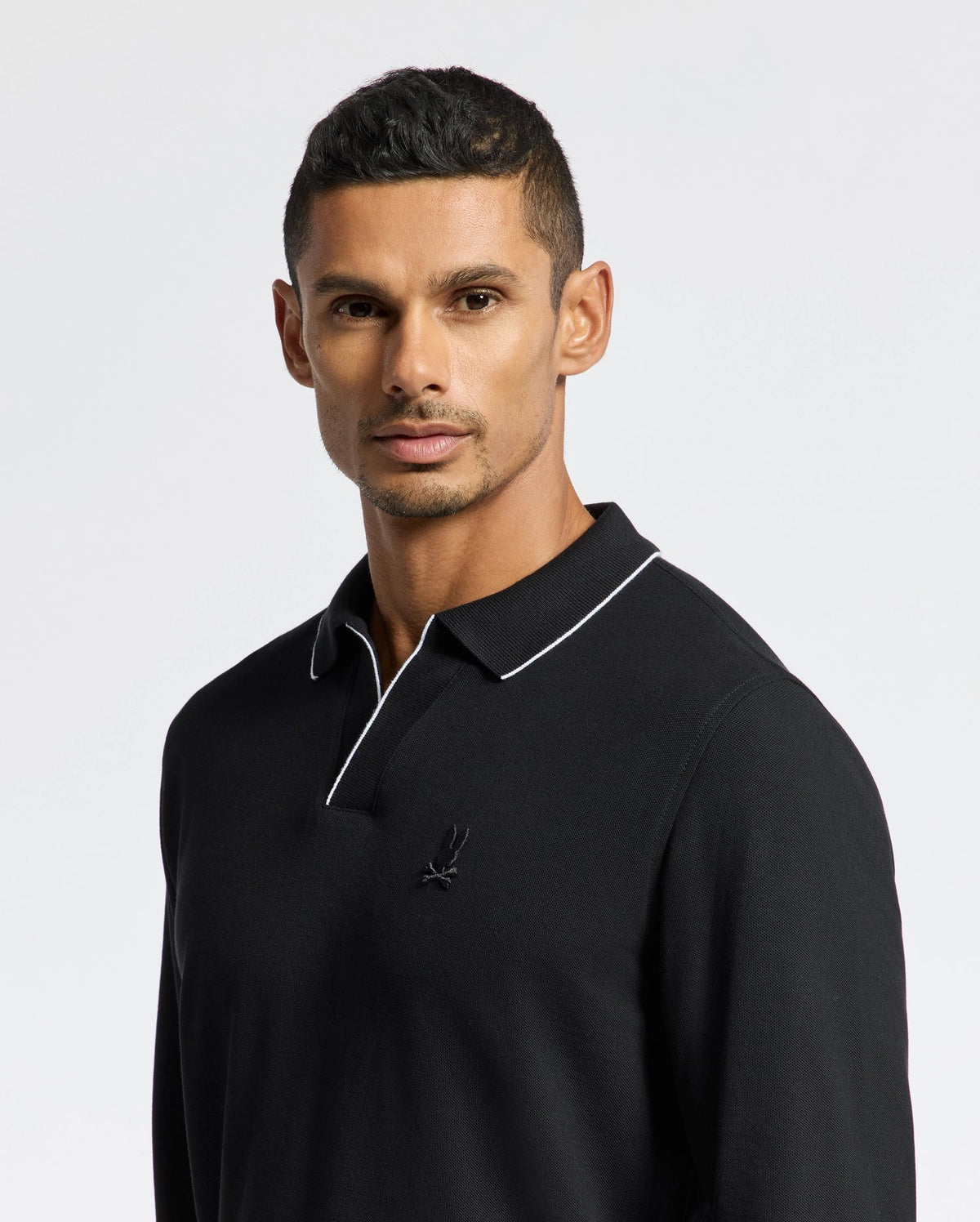 A person with short dark hair and a serious expression stands in front of a plain white background, wearing the MENS CREEDE PIQUE JOHNNY COLLAR POLO from Psycho Bunny. The black long sleeve polo shirt is crafted from soft Pima cotton and features a small embroidered logo along with a white outlined johnny collar.