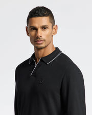 A person with short dark hair and a serious expression stands in front of a plain white background, wearing the MENS CREEDE PIQUE JOHNNY COLLAR POLO from Psycho Bunny. The black long sleeve polo shirt is crafted from soft Pima cotton and features a small embroidered logo along with a white outlined johnny collar.
