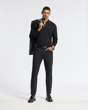 A person stands confidently against a plain background, dressed in a sleek, all-black ensemble. They hold a jacket over their shoulder and wear a watch. Their other hand is in the pocket of their Psycho Bunny Men's Creede Pique Johnny Collar Polo - B6M265E200, and they have short, dark hair.
