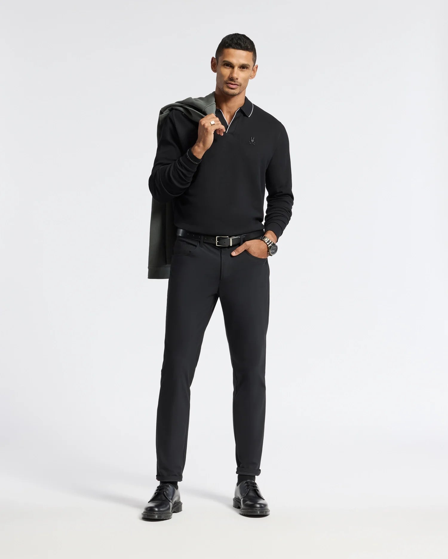 A person stands confidently against a plain background, dressed in a sleek, all-black ensemble. They hold a jacket over their shoulder and wear a watch. Their other hand is in the pocket of their Psycho Bunny Men's Creede Pique Johnny Collar Polo - B6M265E200, and they have short, dark hair.