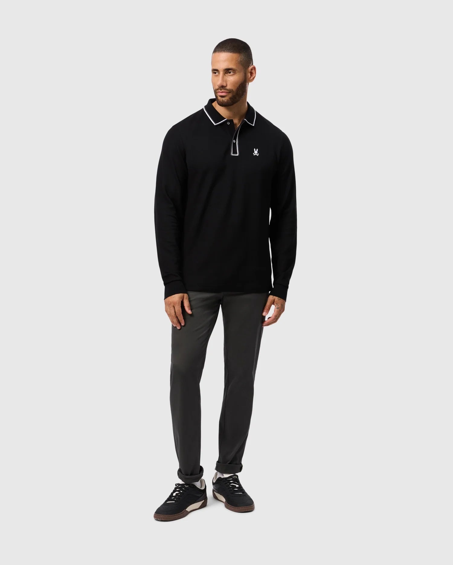A man dressed in the Psycho Bunny MENS KENT LONG SLEEVE PIQUE POLO - B6M935D200, featuring a black color with white trim on the collar, stands against a gray background. His understated elegance is further enhanced by his dark pants and sleek black sneakers.