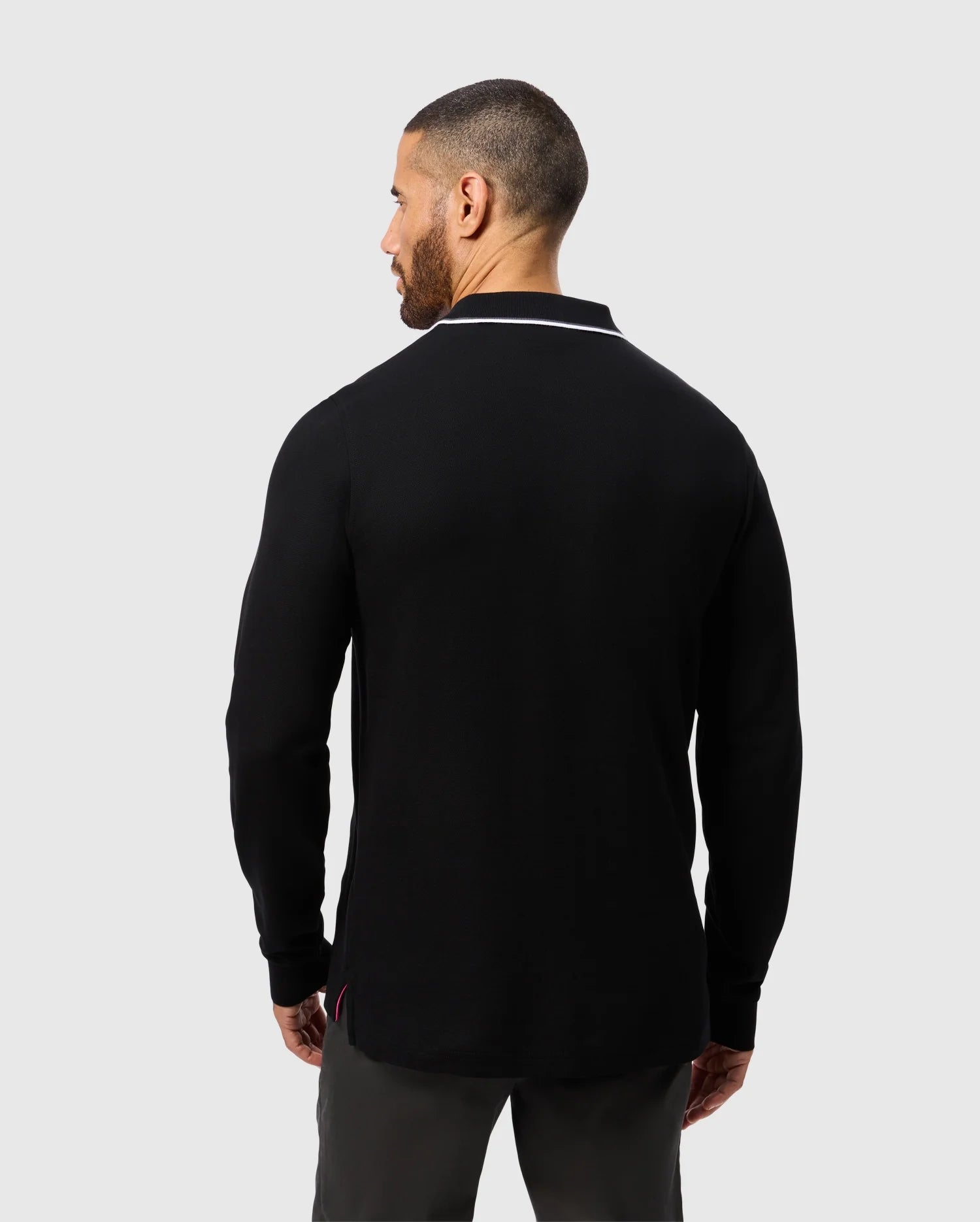 A man with short hair and a beard is facing away, dressed in the Psycho Bunny MENS KENT LONG SLEEVE PIQUE POLO (B6M935D200), made from Pima cotton and highlighted by a white trim on the collar. Combined with dark pants, this outfit emits an understated elegance against the light gray background.