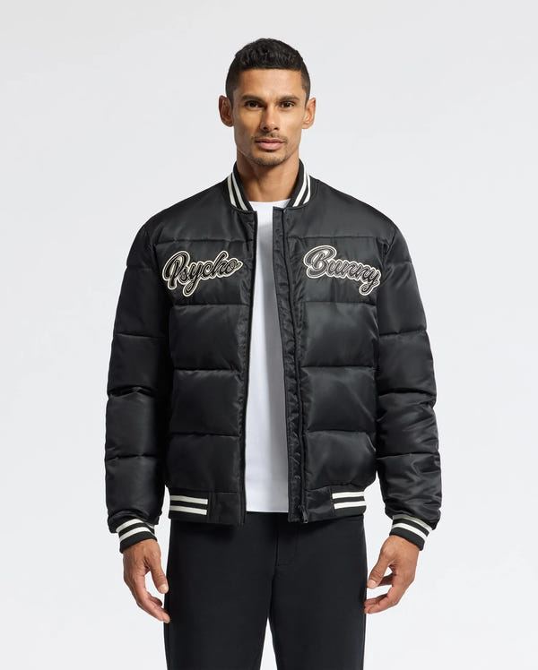 A man in a black MENS FRASER FASHION PUFFER - B6N338E200 by Psycho Bunny stands against a plain background. The lightweight jacket features white striped cuffs and collar, which he pairs stylishly with a white T-shirt underneath and black pants.