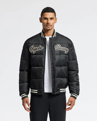 A man in a black MENS FRASER FASHION PUFFER - B6N338E200 by Psycho Bunny stands against a plain background. The lightweight jacket features white striped cuffs and collar, which he pairs stylishly with a white T-shirt underneath and black pants.