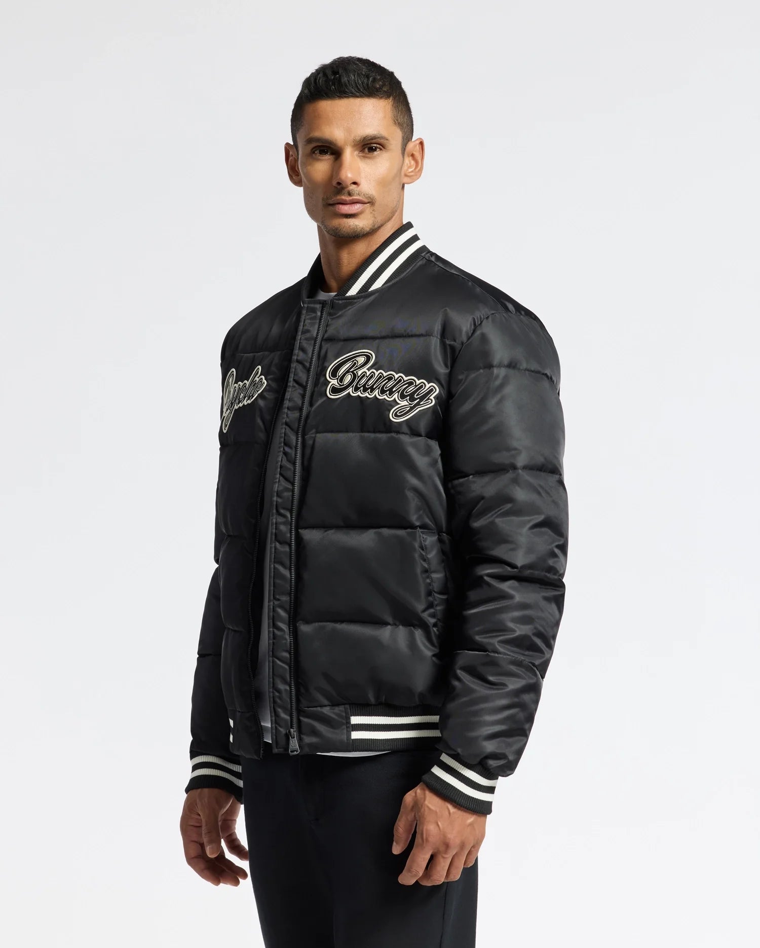 A man wearing a Psycho Bunny MENS FRASER FASHION PUFFER - B6N338E200 in black, featuring white striped cuffs and an embroidered script on the chest. The background is plain white.