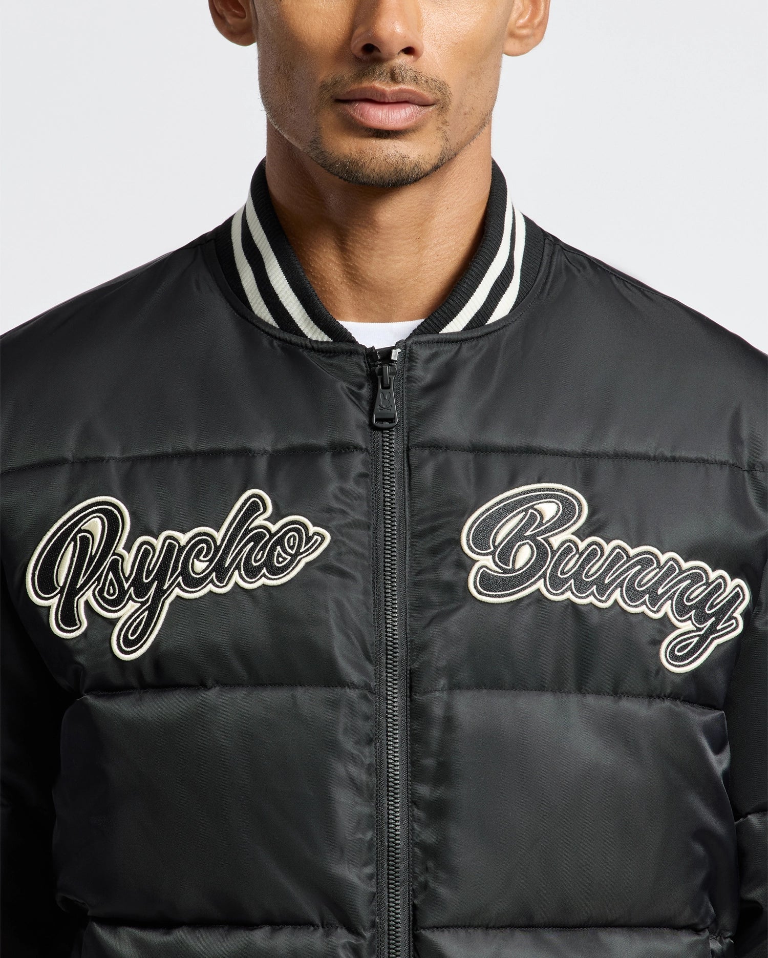 A person is wearing the MENS FRASER FASHION PUFFER - B6N338E200 from Psycho Bunny, featuring a stylish black design with a striped collar and "Psycho Bunny" in white script on the chest. This sleek and lightweight men's puffer coat stands out against a plain white background, perfectly blending style and comfort.
