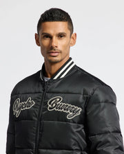 A person wearing a black MENS FRASER FASHION PUFFER - B6N338E200 with the "Psycho Bunny" brand name embroidered on the chest. This stylish jacket, featuring a striped collar, provides warmth in style against a plain white background that highlights this fashionable piece of men's outerwear.
