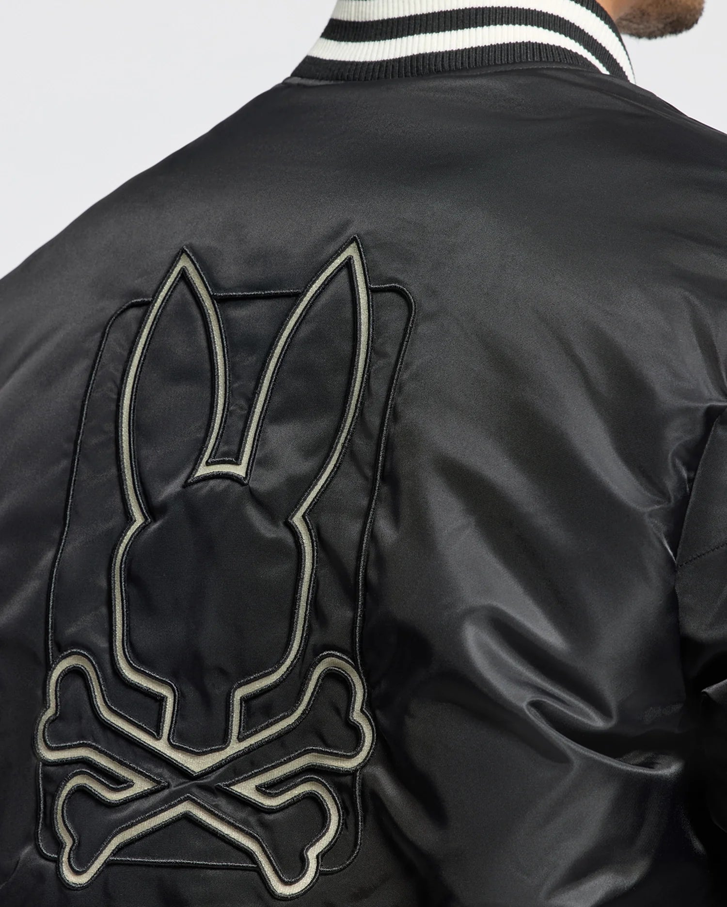 A person is wearing the stylish black MENS FRASER FASHION PUFFER by Psycho Bunny. This warm jacket features a striking large embroidered design on the back, depicting a stylized skull and crossbones with bunny ears. It also boasts a black and white striped collar, effortlessly blending warmth with edgy fashion.