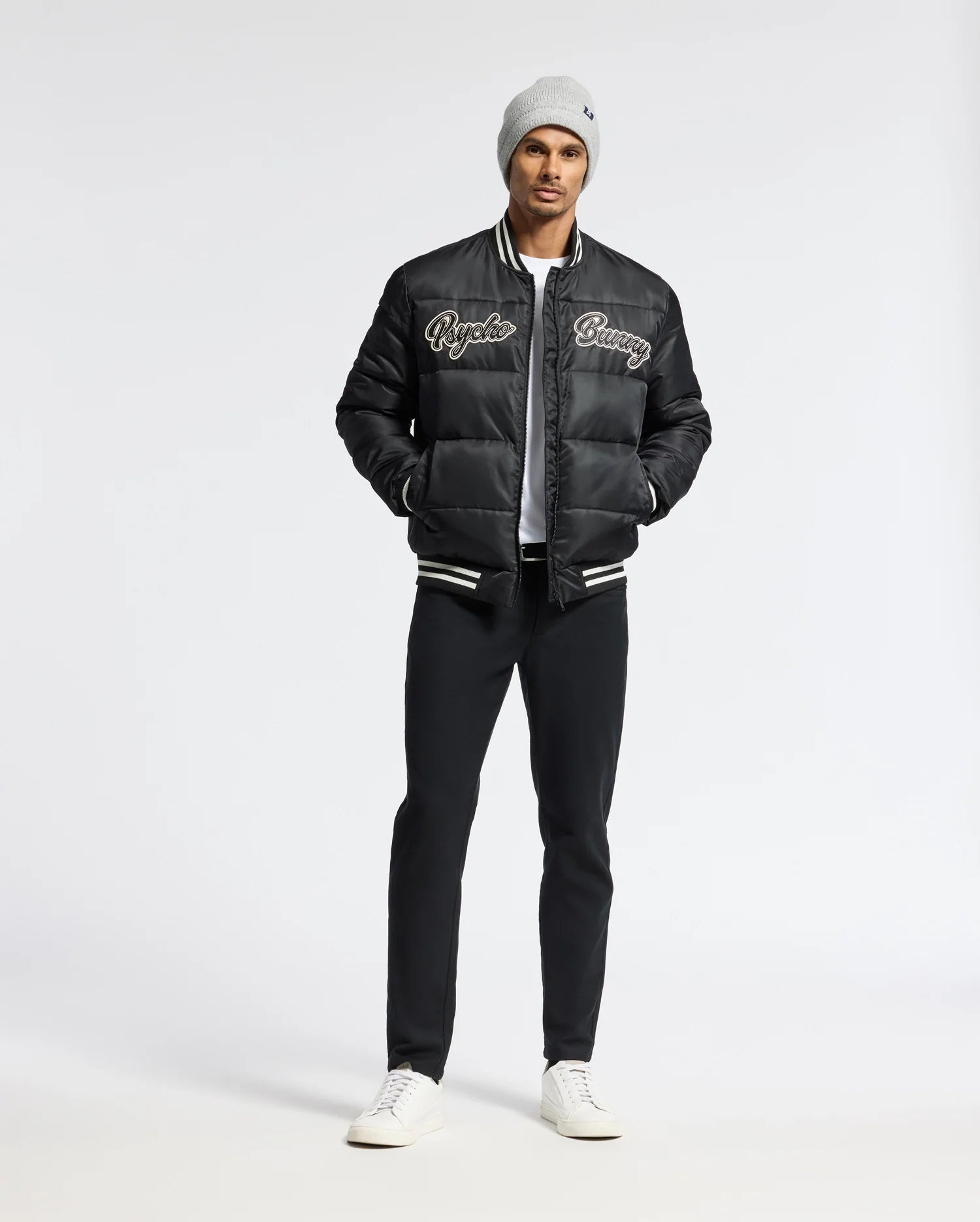 A man wearing a gray beanie and a black bomber jacket with "Richie" and "Savage" text stands in the studio. He showcases a stylish look with the MENS FRASER FASHION PUFFER by Psycho Bunny, paired with black pants and white sneakers, his hands casually tucked into his pockets against a plain backdrop.