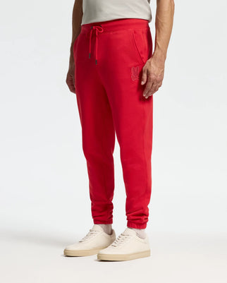 A person is wearing the bright red MENS PIERCE ESSENTIAL FRENCH TERRY SWEATPANT by Psycho Bunny, adorned with a rabbit logo on the pocket. The sweatpants come with an elastic waistband and drawstrings. They pair it with a white shirt and cream sneakers, standing against a plain background.