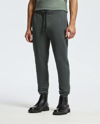 A person stands against a light background wearing the MENS PIERCE ESSENTIAL FRENCH TERRY SWEATPANT - B6P136E200 in dark green by Psycho Bunny, paired with black lace-up boots. The joggers are made from premium cotton and showcase a drawstring waist and a relaxed fit.