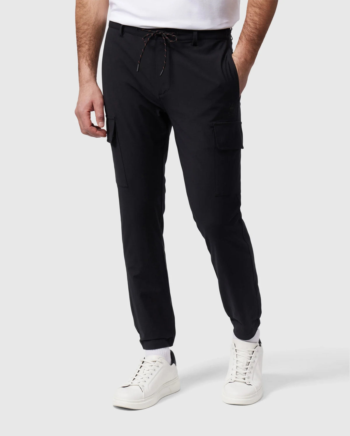 A person is standing, wearing Psycho Bunny's MENS LLOYD CARGO JOGGER - B6P159B200 in black with elasticized cuffs and a white t-shirt. The joggers feature a drawstring closure and utilitarian pockets. They are also sporting white sneakers against a plain light gray background.