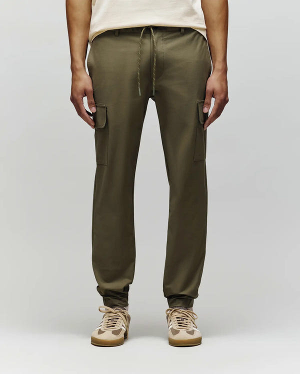 A person in Psycho Bunny MENS LLOYD CARGO JOGGER - B6P159B200, featuring olive green color, drawstrings, and subtle embroidery, pairs them with beige and white sneakers against a plain light gray background.