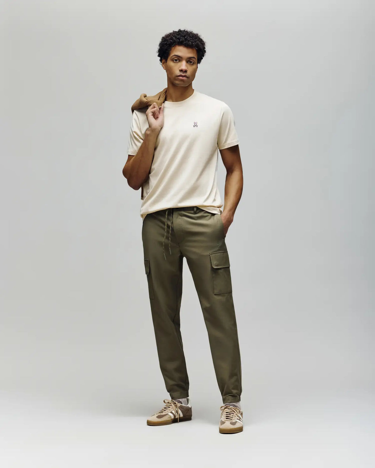 A man stands against a plain background wearing Psycho Bunny's MENS LLOYD CARGO JOGGER - B6P159B200, a beige t-shirt, tan sneakers, and holds a brown jacket over his shoulder, gazing straight ahead with a neutral expression.