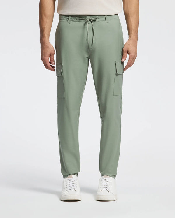 A person models the Psycho Bunny MENS LLOYD CARGO JOGGER - B6P159B200 in sage green, featuring stretch twill, side pockets, and a drawstring waist. Paired with a white shirt and sleek sneakers, the plain background enhances the ensemble's embroidered refinement.