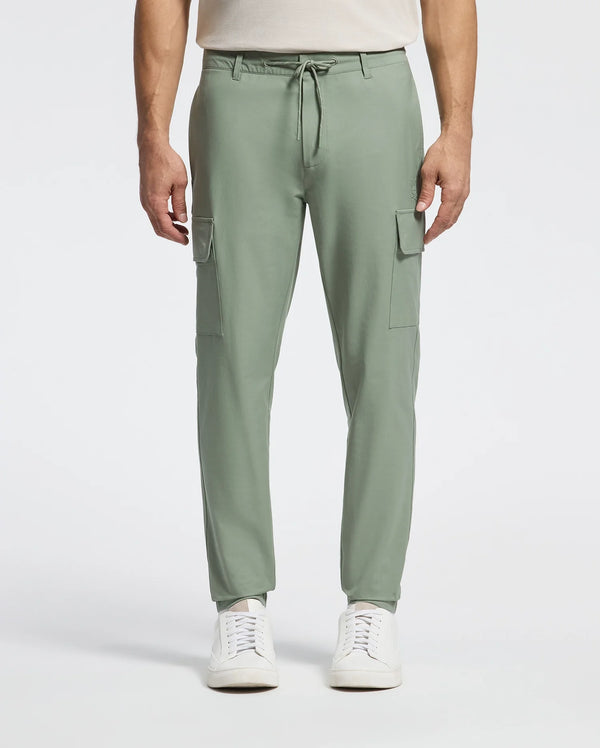A person models the Psycho Bunny MENS LLOYD CARGO JOGGER - B6P159B200 in sage green, featuring stretch twill, side pockets, and a drawstring waist. Paired with a white shirt and sleek sneakers, the plain background enhances the ensemble's embroidered refinement.