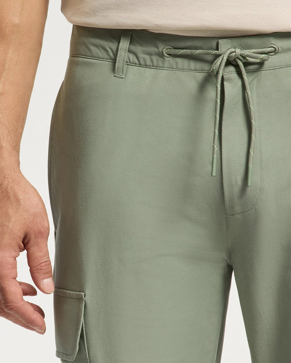 A person is wearing stylish Psycho Bunny MENS LLOYD CARGO JOGGERS in light green with a drawstring waistband. Made from stretch twill, these joggers feature a small side pocket. Their hand, alongside a light-colored shirt, adds embroidered refinement to the ensemble.