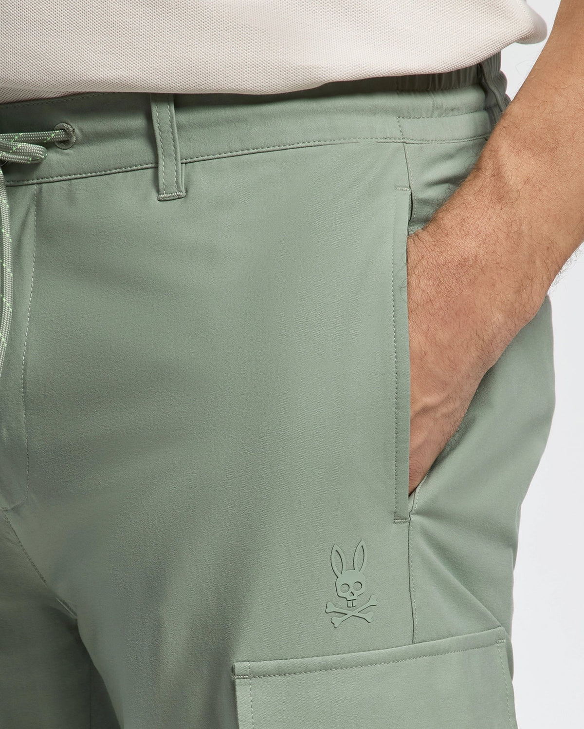 A person wearing Psycho Bunny's Mens Lloyd Cargo Jogger - B6P159B200 in light green stretch twill, featuring an embossed bunny and crossbones logo above the front flap pocket, with one hand in the pocket. They also wear a light-colored shirt.