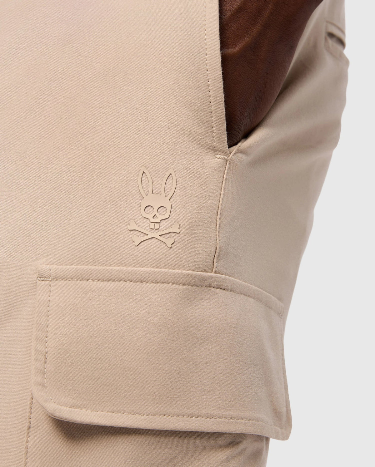 Close-up of a pocket on the MENS LLOYD CARGO JOGGER - B6P159B200 by Psycho Bunny, showcasing a stitched logo of a bunny head with crossbones underneath. The smooth fabric pocket has a flap secured by snap fastening, and a hand is casually resting in the side pocket, highlighting the refined embroidery.