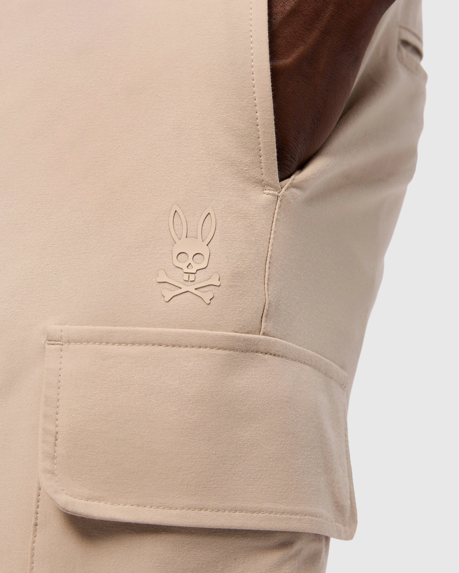 Close-up of a pocket on the MENS LLOYD CARGO JOGGER - B6P159B200 by Psycho Bunny, showcasing a stitched logo of a bunny head with crossbones underneath. The smooth fabric pocket has a flap secured by snap fastening, and a hand is casually resting in the side pocket, highlighting the refined embroidery.