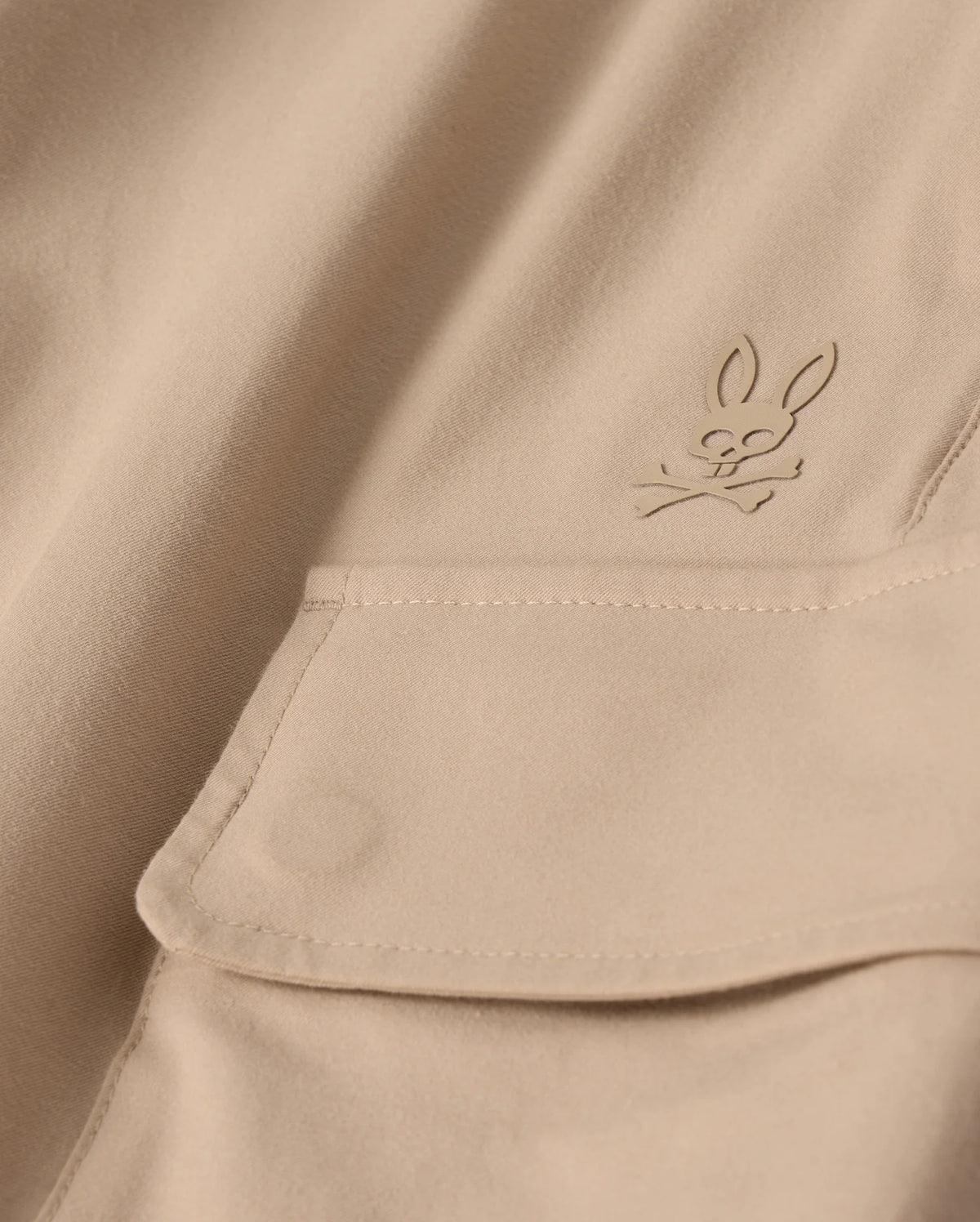 Close-up of the MENS LLOYD CARGO JOGGER - B6P159B200 by Psycho Bunny, featuring a beige fabric adorned with a snap fastening pocket. Above the pocket is a stitched bunny logo with crossed bones underneath its head, showcasing a subtle pirate motif. The playful embroidery refinement adds an elegant touch to the smooth and well-crafted fabric.