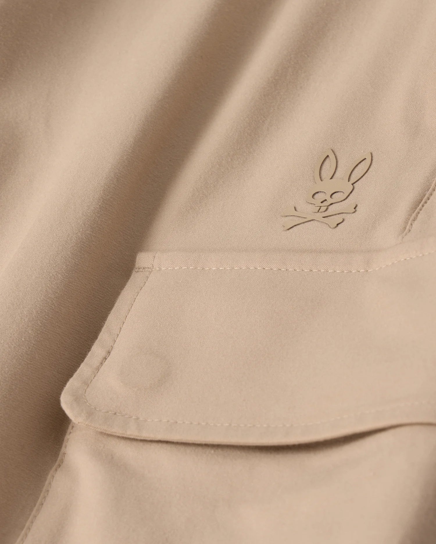 Close-up of the MENS LLOYD CARGO JOGGER - B6P159B200 by Psycho Bunny, featuring a beige fabric adorned with a snap fastening pocket. Above the pocket is a stitched bunny logo with crossed bones underneath its head, showcasing a subtle pirate motif. The playful embroidery refinement adds an elegant touch to the smooth and well-crafted fabric.