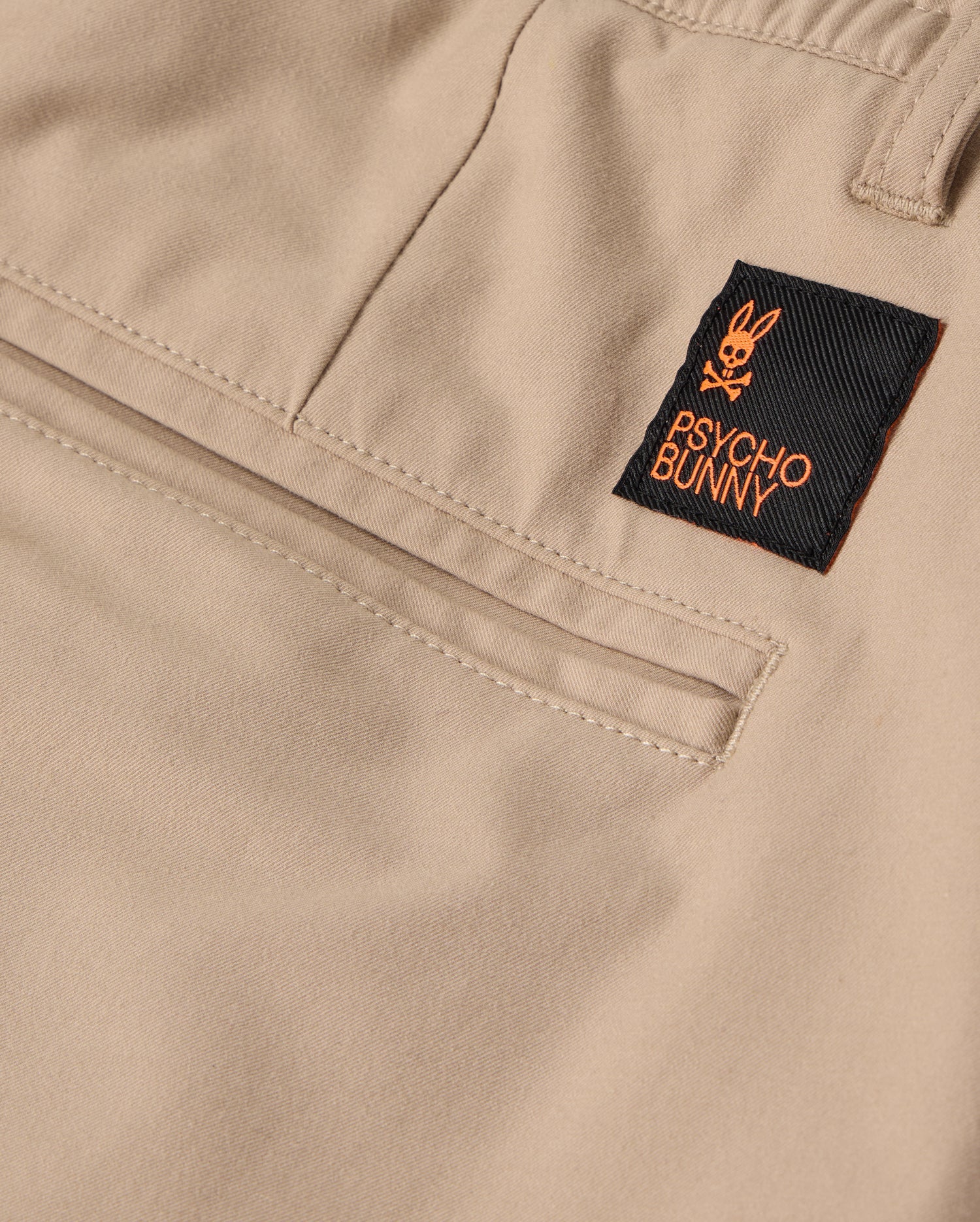 Close-up of the beige fabric MENS LLOYD CARGO JOGGER - B6P159B200 by Psycho Bunny, showcasing an orange-accented black label with a bunny logo and the words "Psycho Bunny" stitched on. The jogger pants exhibit detailed stitching and refined embroidery around the snap fastening pockets.