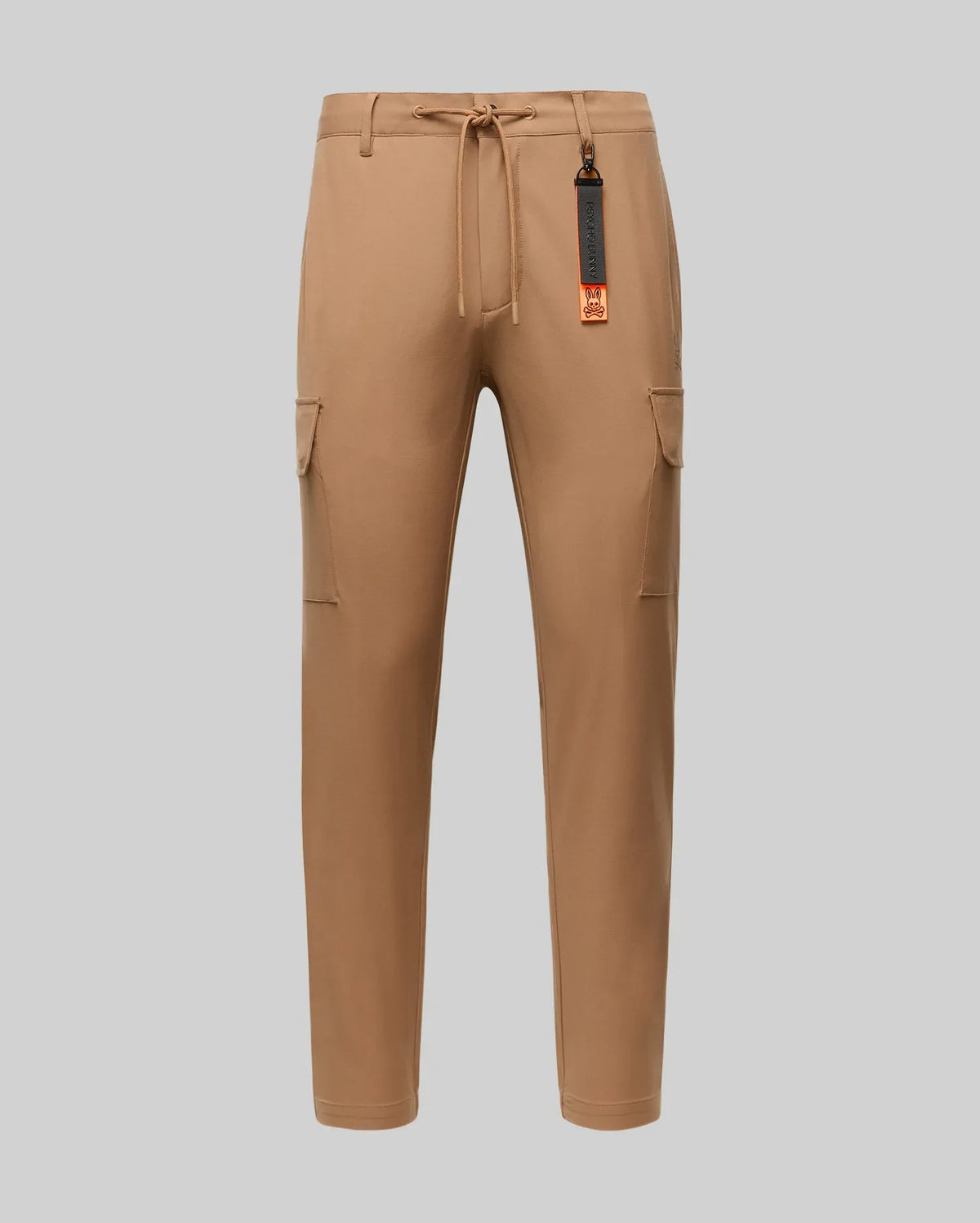 The Psycho Bunny MENS LLOYD CARGO JOGGER - B6P159B200 features tan fabric, elastic waistband, drawstring, side pockets, and a black-and-orange tag on a belt loop—perfect for upgrading your men's pants collection. Shown on a plain light gray background.