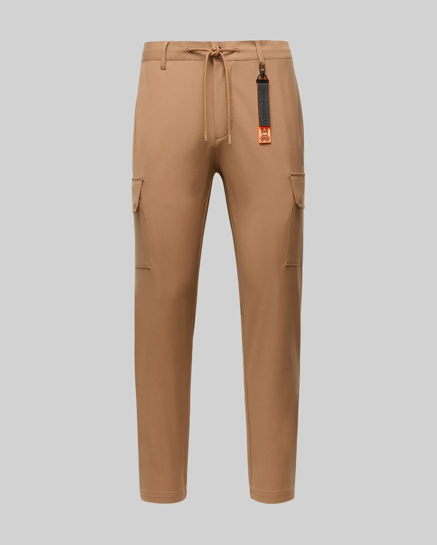 The Psycho Bunny MENS LLOYD CARGO JOGGER - B6P159B200 features tan fabric, elastic waistband, drawstring, side pockets, and a black-and-orange tag on a belt loop—perfect for upgrading your men's pants collection. Shown on a plain light gray background.