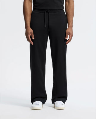 A man is wearing Psycho Bunny's black MENS LYNX WIDE LEG CLASSIC FRENCH TERRY SWEATPANT - B6P379E200 with adjustable drawstring waist, paired with white sneakers, against a plain background. Only his lower body is visible.