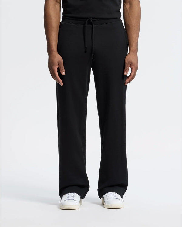 A man is wearing Psycho Bunny's black MENS LYNX WIDE LEG CLASSIC FRENCH TERRY SWEATPANT - B6P379E200 with adjustable drawstring waist, paired with white sneakers, against a plain background. Only his lower body is visible.