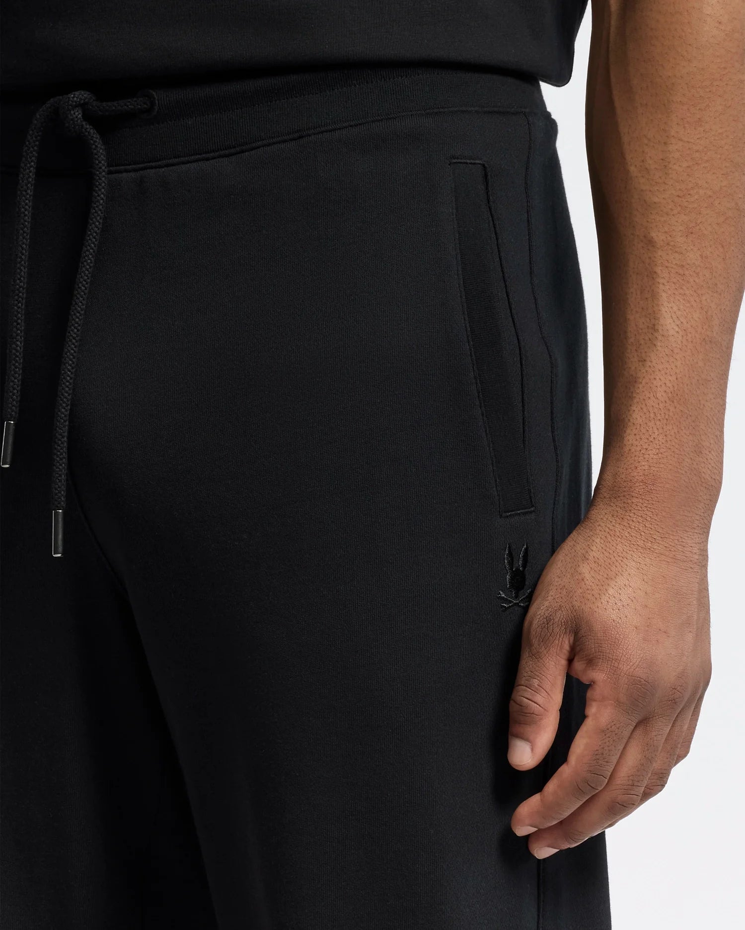 A close-up shows a person in Psycho Bunny's Mens Lynx Wide Leg Classic French Terry Sweatpants (B6P379E200), featuring a drawstring and embroidered logo near the pocket. The individual rests their right hand on their thigh while wearing a matching black shirt.