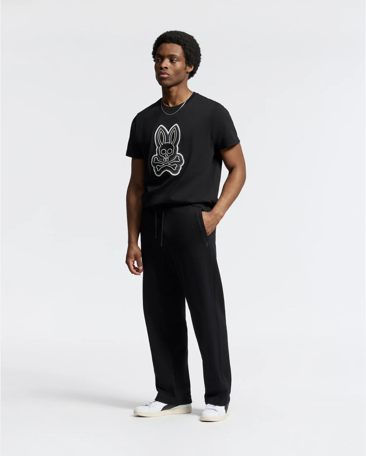 A man wears a Psycho Bunny black T-shirt with a bunny and crossed bones graphic, paired with the MENS LYNX WIDE LEG CLASSIC FRENCH TERRY SWEATPANT and white shoes, standing against a plain background and looking off to the side with a neutral expression.