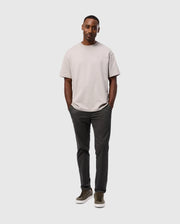 A man confidently models Psycho Bunny's MENS GILMAN CHINO PANT - B6P393Z1WB, effortlessly transitioning from workday to weekend in a light gray T-shirt and chino pants. He pairs them with olive green sneakers, smiling against a white background and epitomizing comfort and style.