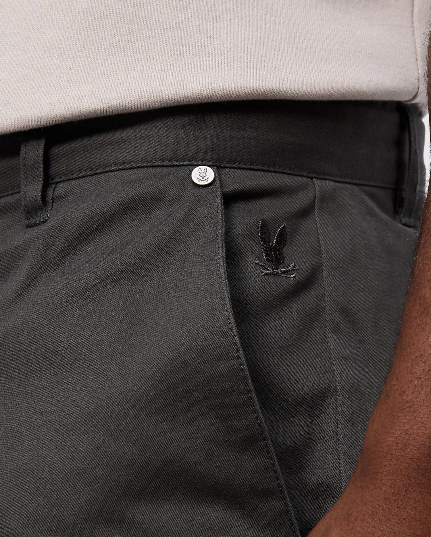 A close-up shows someone in Psycho Bunny's MENS GILMAN CHINO PANT - B6P393Z1WB, dark green with an embroidered bunny logo near the front pocket. The comfort suits workdays to weekends, paired with a tucked-in light shirt. A logo metal button is visible on the waistband.