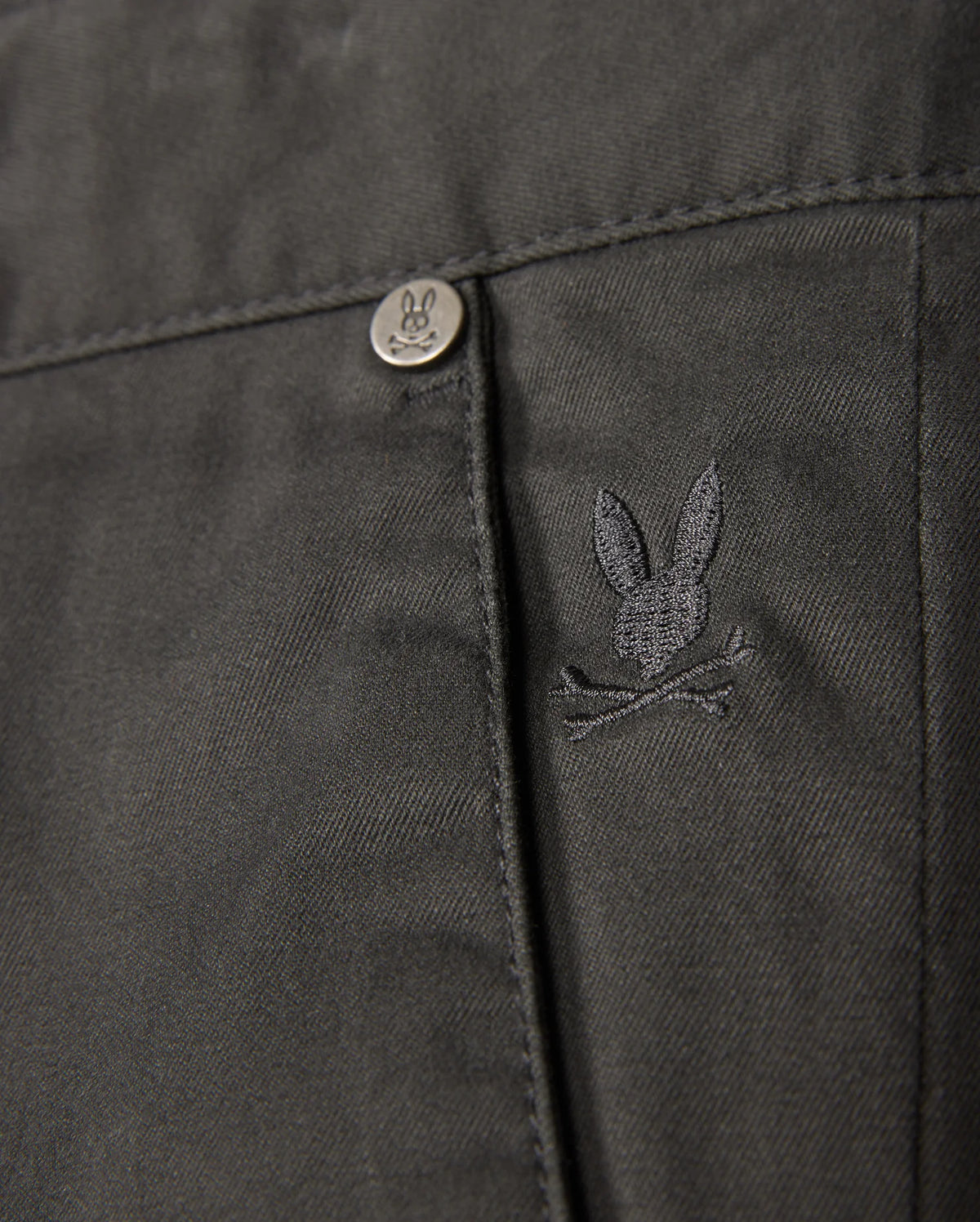 The Psycho Bunny MENS GILMAN CHINO PANT - B6P393Z1WB features a close-up of fabric with an embroidered rabbit head and crossed bones logo. A metal button displays the same design. This dark, comfy chino transitions smoothly from workday to weekend, making it a versatile wardrobe staple.