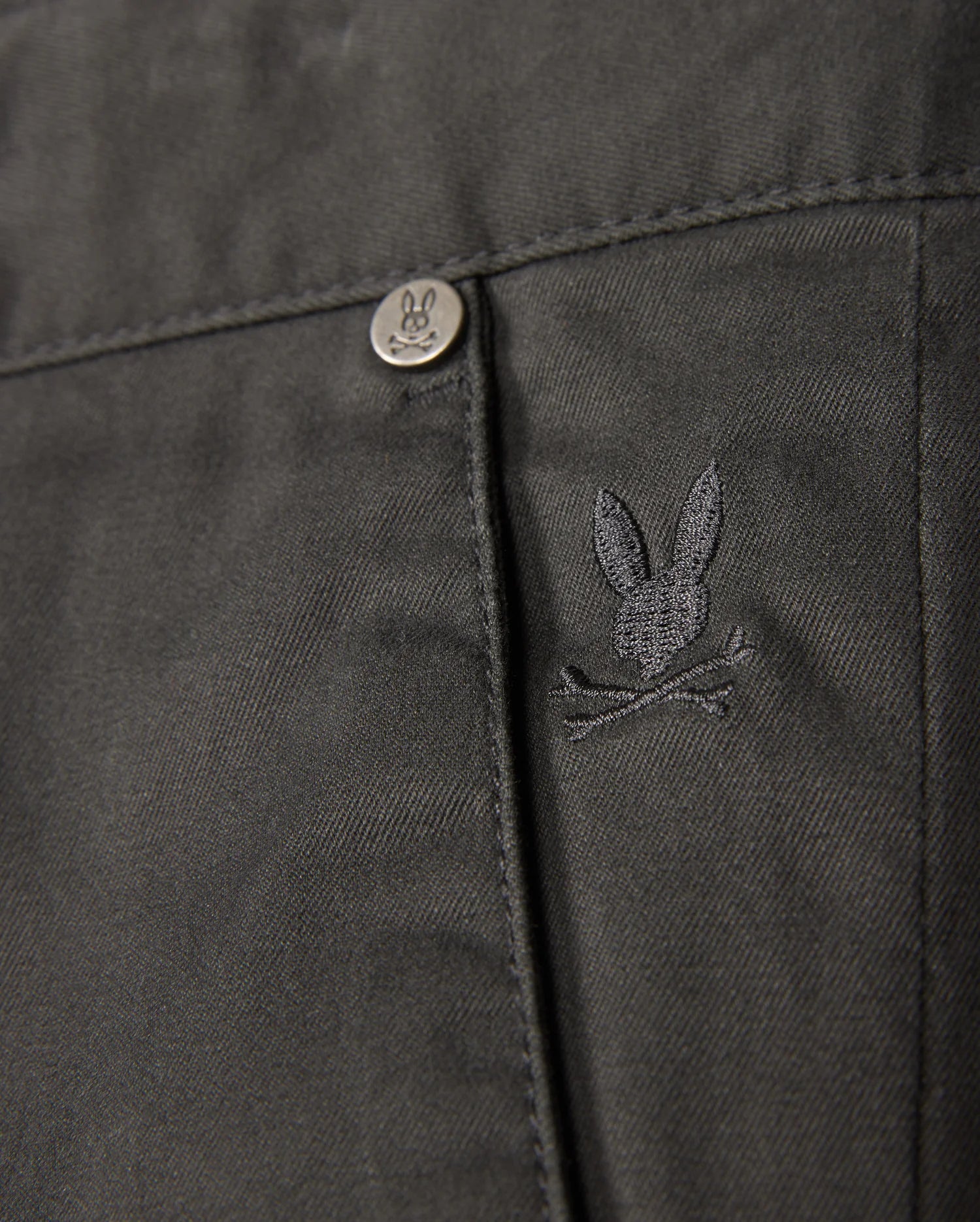 The Psycho Bunny MENS GILMAN CHINO PANT - B6P393Z1WB features a close-up of fabric with an embroidered rabbit head and crossed bones logo. A metal button displays the same design. This dark, comfy chino transitions smoothly from workday to weekend, making it a versatile wardrobe staple.