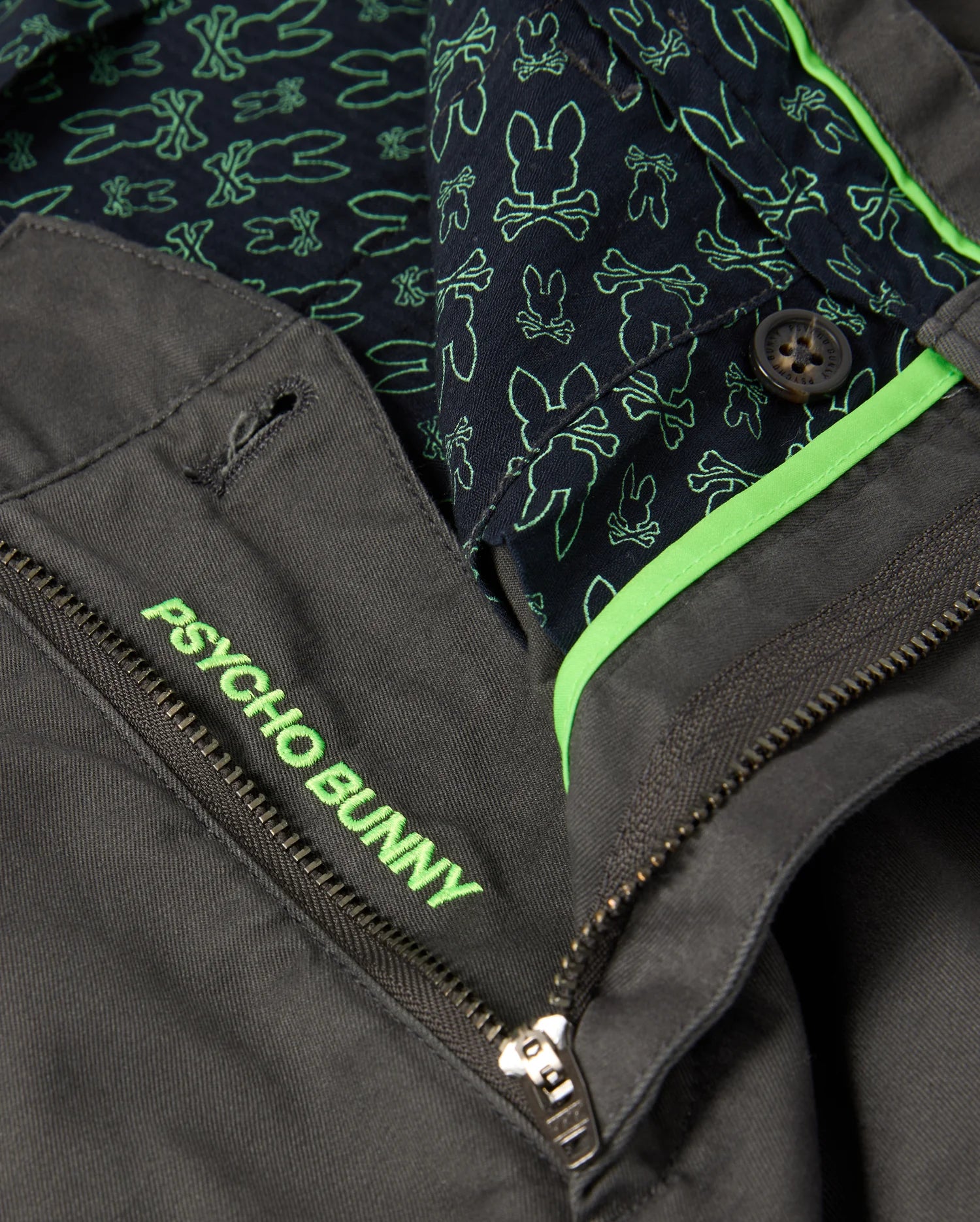 Close-up of the MENS GILMAN CHINO PANT - B6P393Z1WB by Psycho Bunny in dark gray, featuring "Psycho Bunny" embroidered in neon green on the pocket. Designed for comfort from workday to weekend, with an inner lining of outlined bunnies and a partially open zipper showing more green accents.