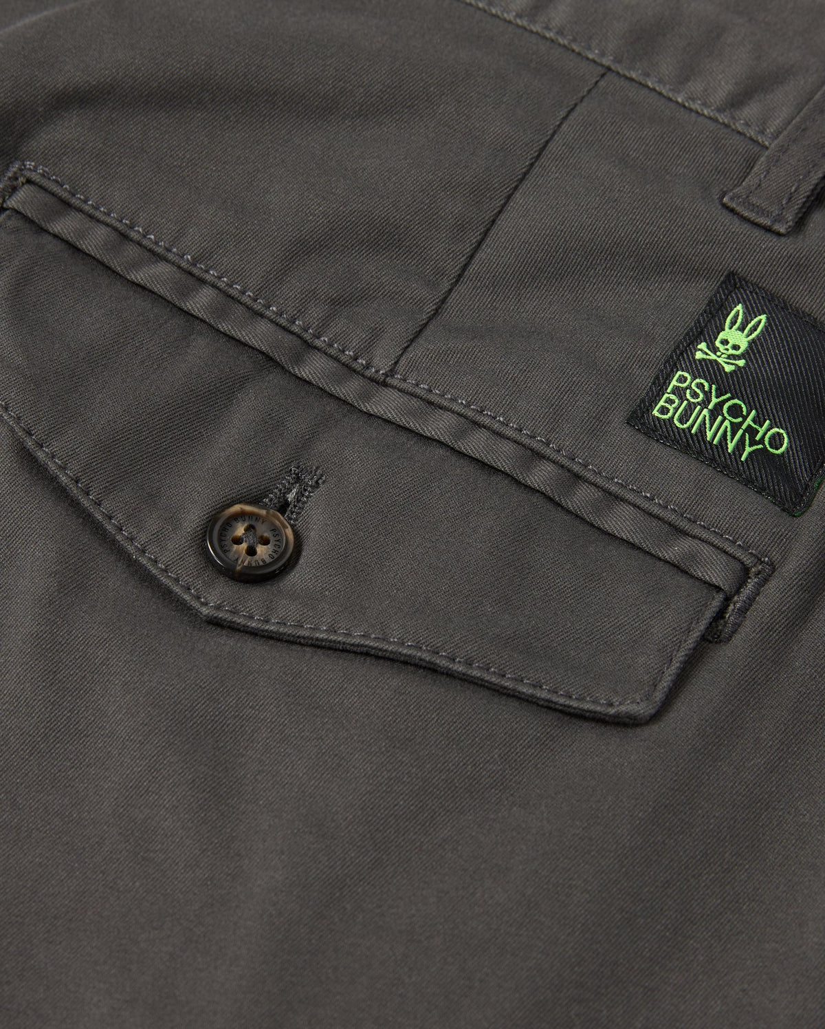 Close-up of a dark gray shirt pocket with button fastening. Above it, a black and green label features a bunny design and "Psycho Bunny." Pairing perfectly with the MENS GILMAN CHINO PANT - B6P393Z1WB from Psycho Bunny, this classic style smoothly transitions from workday to weekend.
