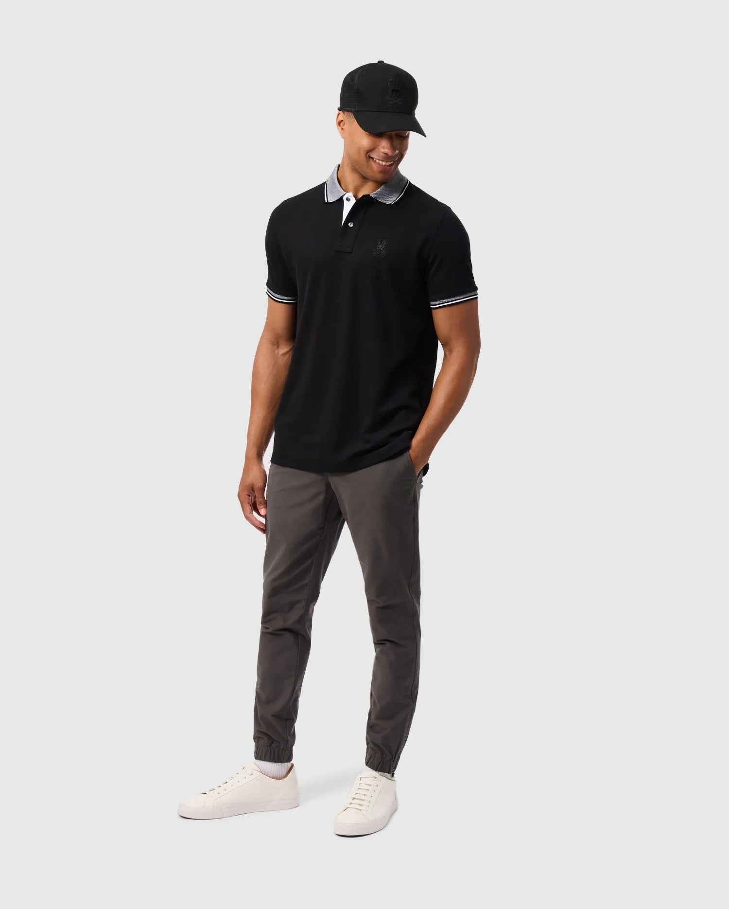 A man is standing and smiling, dressed casually in a black polo shirt, grey Psycho Bunny MENS MADISON COMMUTER PANT - B6P395Z1WB, white sneakers, and a black cap. He is looking down with one hand in his pocket that holds a detachable Bunny keychain. The background is plain and light grey.