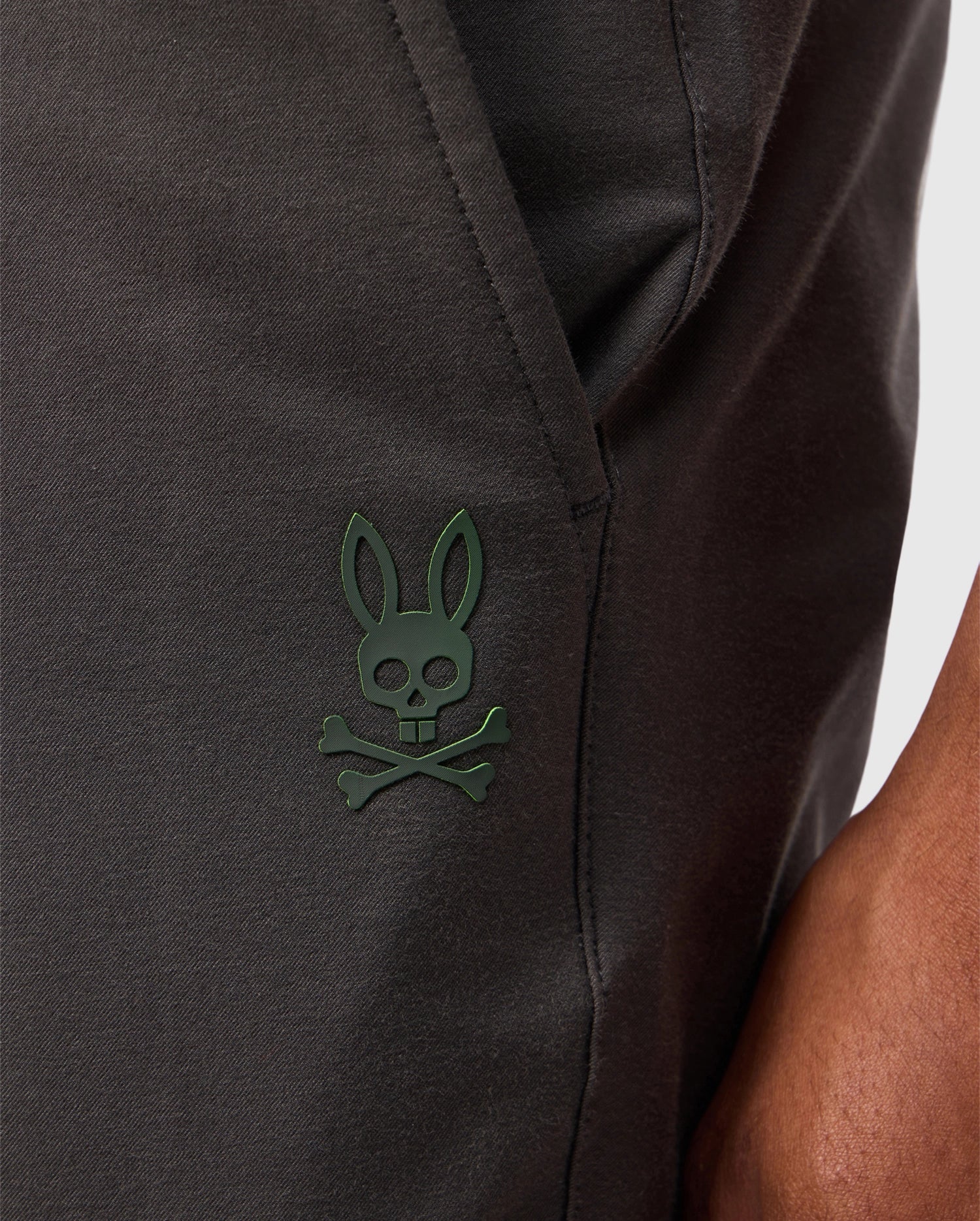 Close-up of a person wearing dark-colored Psycho Bunny MENS MADISON COMMUTER PANT - B6P395Z1WB. The pants feature a small green skull and crossbones logo with bunny ears near the pocket, showcasing subtle Bunny detailing. The person's arm is partially visible on the right side of the image.